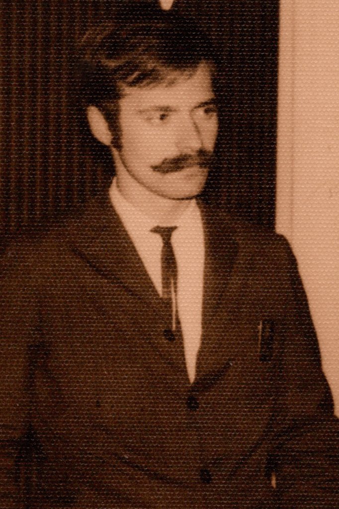 Foto em preto e branco em tons amarelados, impressa em papel com textura, de um homem com expressão séria. Ele tem aproximadamente 40 anos, cabelos curtos penteados para o lado, olha em diagonal para a direita, tem bigode, usa terno preto com gravata fina preta e camisa branca. Está enquadrado até a altura da barriga.
