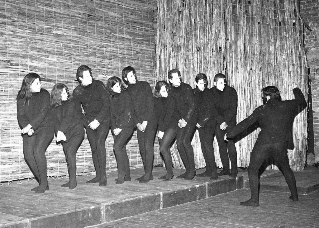 Foto em preto e branco de uma cena teatral. Um grupo de 9 pessoas está em fila, em cima de um tablado. Todos vestem blusa, calça e meias de malha pretas e estão com os rostos pintados, a metade direita de branco e a metade esquerda de preto. Eles tem o tronco ligeiramente voltado para frente, as mãos unidas na altura da cintura e olham para o lado direito, com expressão séria, para um outro personagem, vestido da mesma forma, que está em frente ao tablado, de costas para nós, com a mão direita levantada, como se fosse golpear os outros com um chicote. O cenário ao fundo é de paredes feitas de grandes esteiras de palha.
