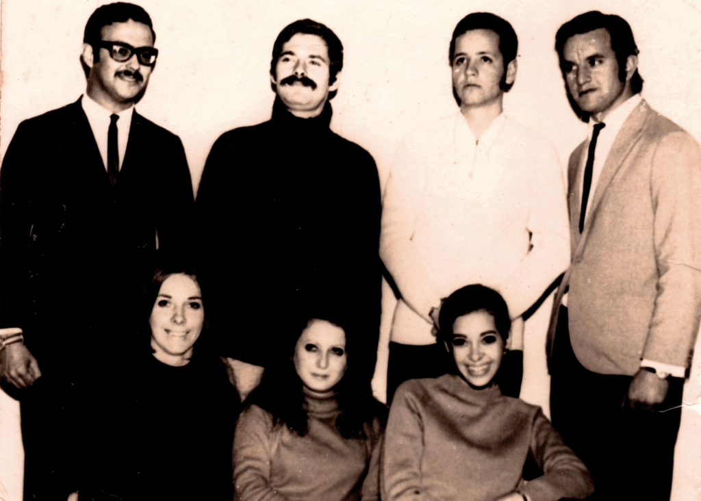 Foto em preto e branco de quatro homens em pé perfilados e três mulheres sentadas. Os quatro homens olham para diferentes pontos, tem cabelos curtos e escuros. As três mulheres olham para nós. O homem bem da esquerda usa óculos com armação grossa preta, bigode, terno preto com gravata fina preta e camisa branca. Faz um leve sorriso e tem o punho da mão direita cerrado. Ao lado dele, o segundo homem usa blusão preto com gola alta, bigode e olha para o longe, com expressão serena. O terceiro homem olha para o alto com expressão séria, tem suíças e usa casaco branco. O homem da direita está ligeiramente de perfil esquerdo, cenho franzido, usa suíças, blazer bege, gravata fina preta, camisa branca e parece estar com o punho da mão esquerda cerrado. As mulheres da esquerda e da direita sorriem, enquanto a do centro está séria, com olhar penetrante. A da esquerda tem cabelos e roupa pretos, que se confundem com as roupas dos homens que estão atrás dela. A do centro tem cabelos pretos e lisos abaixo dos ombros e usa blusa em tom cinza escuro. A da direita tem cabelos presos para trás e usa blusa em tom cinza escuro. As mulheres estão enquadradas até a altura da barriga. O fundo é uma parede branca.
