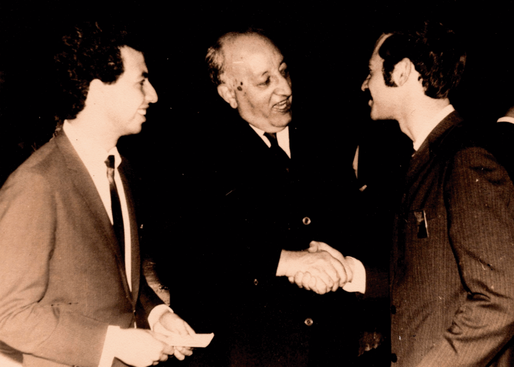 Foto em preto e branco de três homens em um momento descontraído. O da esquerda está de perfil direito, sorri e olha para os outros dois, que se cumprimentam. Os três usam roupas formais. O do centro é um senhor, com terno preto e camisa branca. Ele cumprimenta e conversa com um homem que está à direita, de perfil esquerdo, parcialmente de costas para nós e usa terno com listras discretas.
