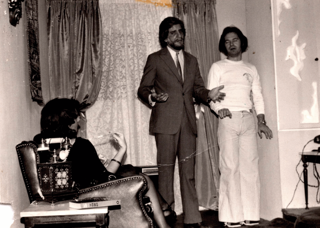 Foto em preto e branco de uma cena de teatro ou filme em uma sala. Dois homens estão em pé, ao centro. Um deles parece argumentar, com as palmas das mãos voltadas para o alto. Ele usa terno e gravada. Ao lado dele, à direita, um homem com cabelo Chanel, blusa de manga comprida branca e justa e calça boca de sino quadriculada em tons claros. À esquerda, uma pessoa que parece ser uma mulher está sentada em uma poltrona de couro, de costas para nós e olha para eles. Uma pequena mesa de canto com um livro e um telefone antigo está próxima a ela. Ao fundo, uma janela com cortinas antigas em duas camadas.
