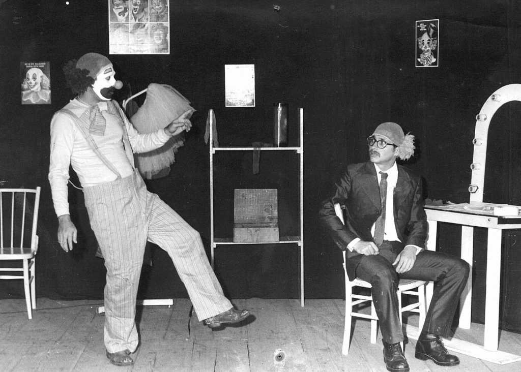 Foto em preto e branco em um teatro. À esquerda, um palhaço com o pé esquerdo um pouco levantado e o tronco levemente inclinado para trás, de perfil direito. Ele aponta e fala com outro homem, que está sentado à direita, em uma cadeira. O palhaço usa touca que simula como se ele fosse calvo na parte de cima da cabeça e tivesse cabelos volumosos escuros na parte de trás. Tem nariz de palhaço, rosto pintado de branco com contorno preto grosso em volta da boca, blusa branca de mangas compridas, suspensório e calça folgada com listras verticais em tons de cinza. O homem que está sentado usa touca semelhante, com cabelos claros na parte de trás, óculos com armação preta, terno preto com camisa branca, gravata preta e botinas pretas de couro.  Ao fundo, uma cortina preta de teatro, uma cadeira à esquerda, uma pequena estante ao centro, quadros com desenhos de palhaços e, à direita, próximo ao homem sentado, uma bancada com espelho e luzes, como um camarim.
