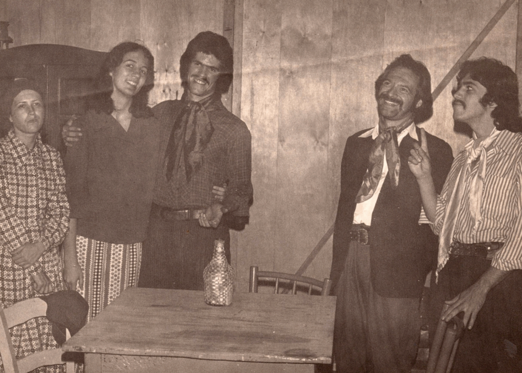 Foto em preto e branco de um grupo de 5 pessoas que estão em pé, três à esquerda e duas à direita, com figurinos tradicionais gaúchos. Uma mesa simples de madeira está ao centro, com uma garrafa apoiada no tampo. À esquerda, uma mulher de baixa estatura, expressão enfadonha, lenço na cabeça e vestido estampado, com gola alta e mangas compridas. Ao lado dela, uma mulher jovem e um homem abraçados e sorridentes. Ela tem cabelos levemente ondulados na altura dos ombros, usa blusa escura de manga comprida e saia longa estampada. Ele tem cabelos volumosos que cobrem as orelhas, usa lenço no pescoço, camisa xadrez escura para dentro da bombacha e cinto de couro. À direita, dois homens, um deles que sorri e outro que fala algo com o dedo em riste. Ambos usam lenço no pescoço, o primeiro tem bigode, blazer escuro, camisa branca para dentro da bombacha e cinto de couro com fivela de metal. O da direita usa bigode fino, camisa listrada na vertical em tons contrastantes para dentro da bombacha e cinto de couro.
