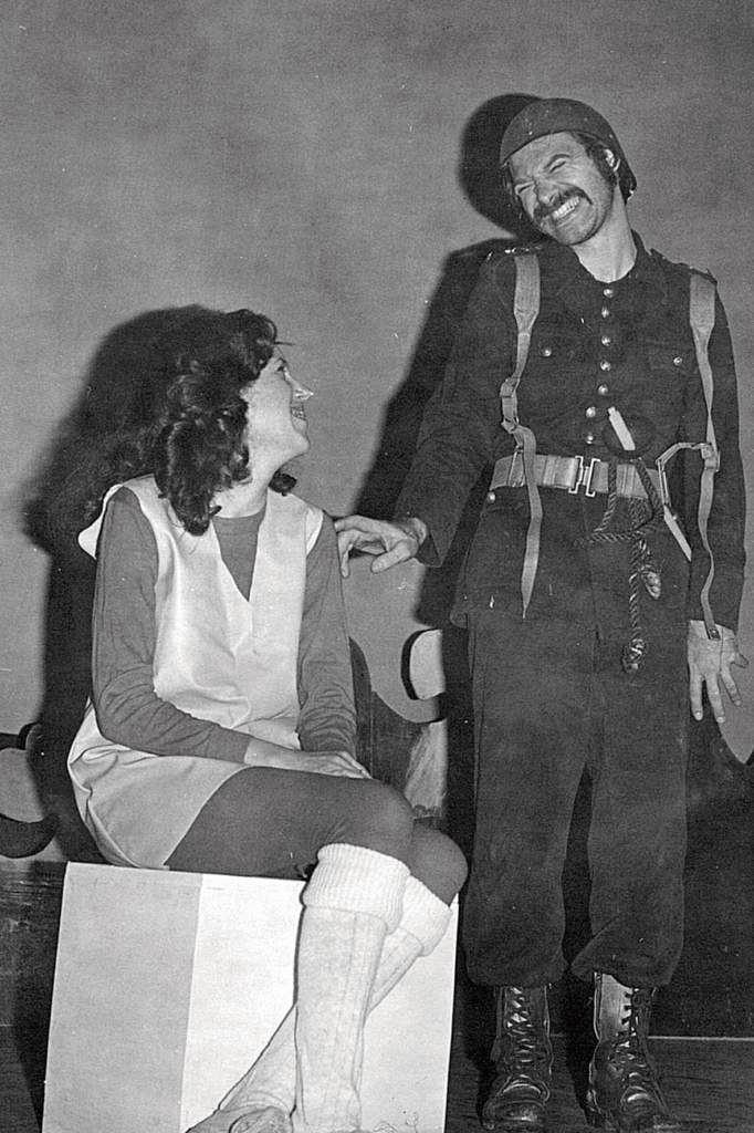 Foto em preto e branco de uma cena teatral do mesmo espetáculo da imagem anterior. A mulher está sentada no cubo branco e olha para o homem de roupa preta, que olha para ela, toca em seu ombro e faz um grande sorriso forçado. A mulher usa meia três quartos branca. O homem usa capacete e roupa militar preta, com um cinto e suspensório de couro. 
