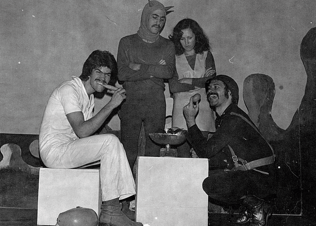 Foto em preto e branco de uma cena teatral com quatro personagens, um sentado à esquerda, de perfil direito, dois em pé ao centro de frente para nós e um agachado à direita, de perfil esquerdo. O da esquerda, que usa roupas brancas, morde um objeto que parece um sacolé, com o rosto contraído, como se fizesse força. Ao centro, um homem e uma mulher estão com os braços cruzados e olham para o homem da direita, que sorri com um ovo na mão, próximo ao seu rosto. Ele está com o cotovelo apoiado em um pequeno cubo branco, que está no centro da cena. Usa chapéu, roupas e coturnos pretos. O homem que está em pé usa uma touca com dois pequenos chifres, como um diabo. A mulher usa um vestido sobre blusa de manga comprida e calça.
