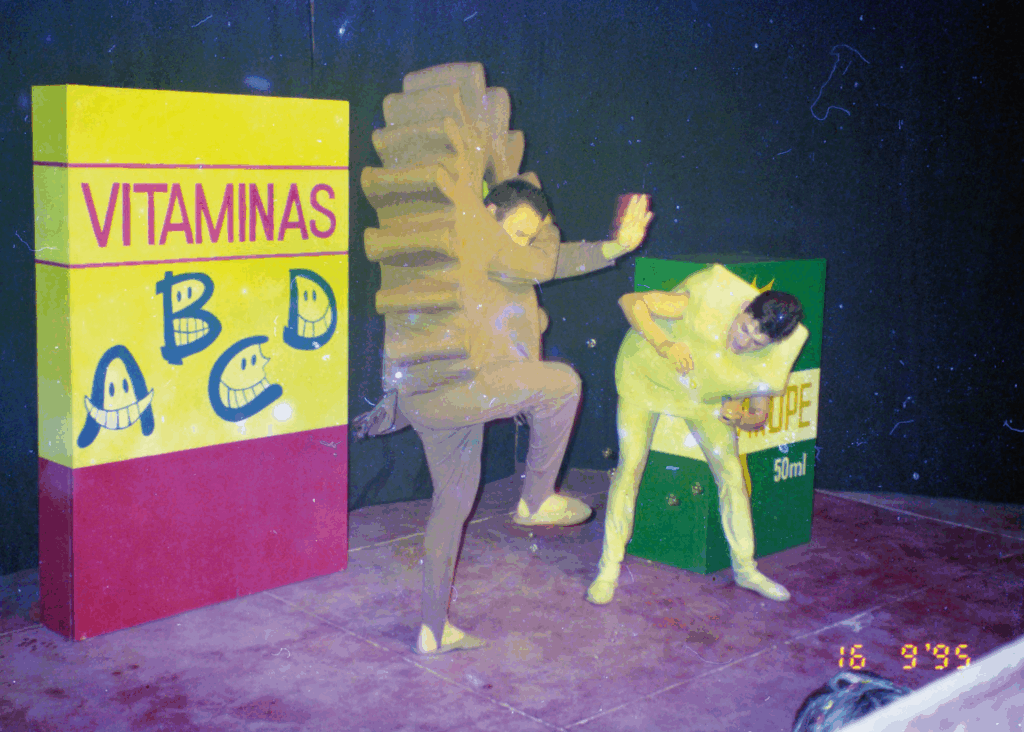Foto colorida de uma cena de teatro infantil. À esquerda, uma grande caixa cenográfica de vitaminas A, B, C e D. As letras são desenhos com olhos e bocas, como se fossem rostos que sorriem. Ao centro, dois personagens em movimento de luta. Um deles tem figurino marrom semelhante a uma engrenagem, como se fosse um vírus ou bactéria. Ele está com a perna direita levantada, o braço esquerdo esticado para frente e a mão direita protegendo o rosto. O outro personagem tem um figurino amarelo claro que parece uma caixa retangular. Ele tem o tronco inclinado para frente e segura um brinquedo de bolinhas de sabão. Algumas bolinhas flutuam pelo ar e o primeiro personagem parece se defender delas. Atrás do segundo personagem, há uma grande caixa cenográfica escrito xarope.
