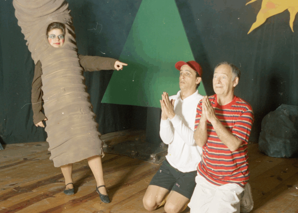 Foto colorida de cena de teatro infantil. À esquerda, uma personagem feminina, com um figurino marrom sanfonado, como uma lombriga. À direita, dois homens estão ajoelhados com as mãos unidas em frente ao peito, como se rezassem. Um deles é jovem e o outro é um senhor. Ambos usam roupas casuais. Ao fundo, uma árvore cenográfica.
