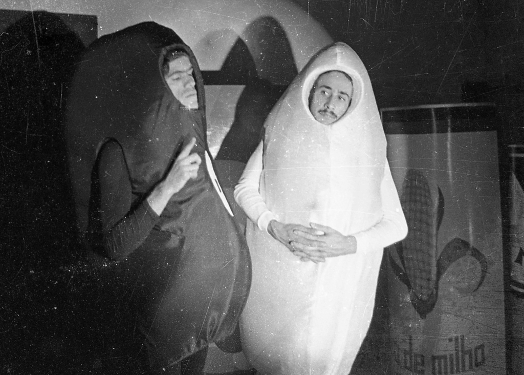 Foto em preto e branco, com sinais de desgaste do tempo como pontinhos e riscos finos brancos. Em uma cena teatral, dois personagens conversam. Eles tem figurinos grandes e ovalados, onde só aparecem seus rostos. Um deles é escuro e outro é claro.  Ao fundo, uma grande lata cenográfica de milho em conserva.
