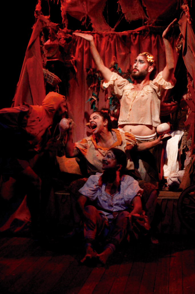 Foto colorida com iluminação tênue avermelhada de uma trupe teatral em cena. Ao centro, uma mulher está sentada no chão do palco. Ela sorri e olha para um personagem que está à esquerda, em pé e com o tronco inclinado na direção de outra mulher, que sorri para ele e está posicionada logo atrás da primeira mulher. Atrás deles, um homem está em pé, com as mãos levantadas para o alto. Todos tem figurinos excêntricos, alguns tem os rostos pintados e o cenário ao fundo é uma estrutura com tecidos em farrapos vermelhos.