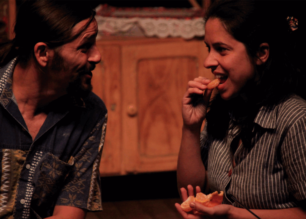 Foto colorida de uma cena teatral. Um homem fala algo com as sobrancelhas levantadas para uma jovem, que está prestes a morder um gomo de bergamota. Ele tem cabelos pretos e compridos presos para trás e usa uma camisa azul com estampas brancas e amarelas escuras. A jovem tem cabelos pretos volumosos na altura dos ombros e usa uma camisa cinza escuro com listras finas brancas. Eles estão enquadrados da barriga para cima.
