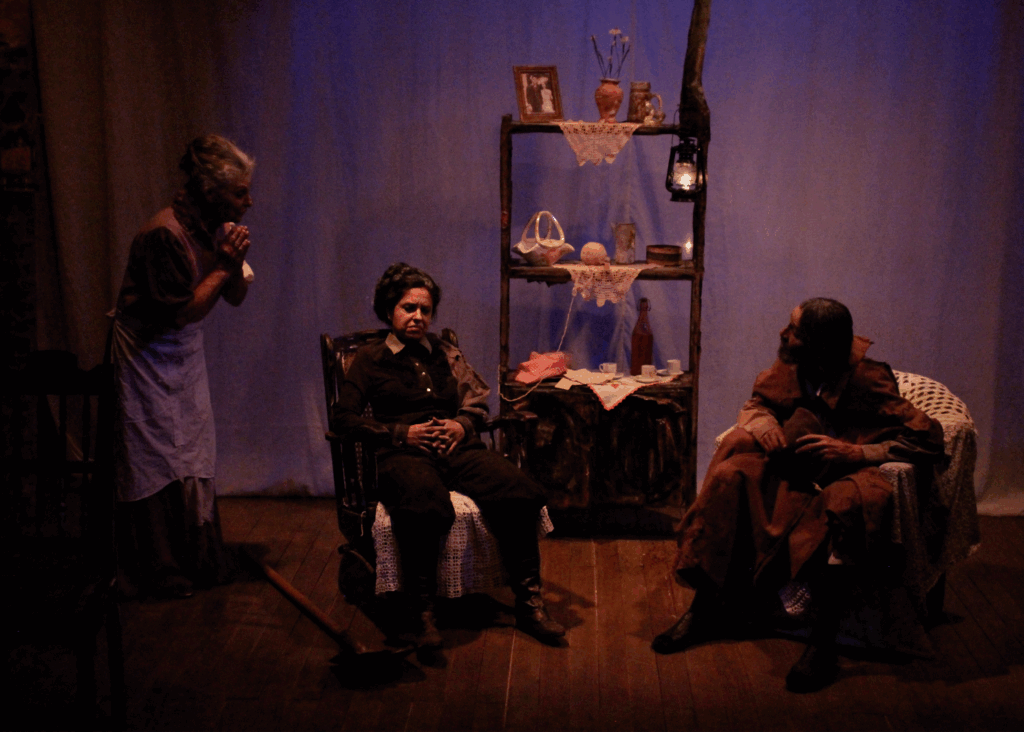 Foto colorida com luz tênue de uma cena teatral. À esquerda, uma senhora de avental branco, mãos unidas abaixo do queixo e o tronco levemente inclinado para frente. Ela parece falar algo. Ao centro, outra senhora sentada em uma cadeira de balanço antiga de madeira. Ela tem os cabelos presos em coque, as mãos unidas com os dedos entrelaçados em frente à barriga, roupas escuras e está séria. À direita, um senhor de cabelos compridos, sentado em um sofá de um lugar, com um grande sobretudo marrom, olha para a primeira senhora. Ao fundo, uma estante de madeira com objetos como porta retrato, vaso, caneco e lampião na parte de cima, novelo de lã, porcelanas, xícaras de café e outros objetos mais abaixo.
